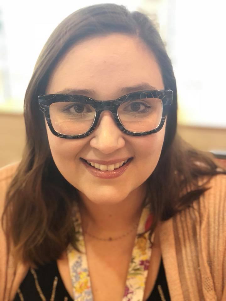 A woman smiling at the camera, wearing dark-framed glasses and a light sweater